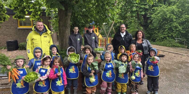 Kinder stehen vor einem Baum und halten Gemüse in Richtung Kamera