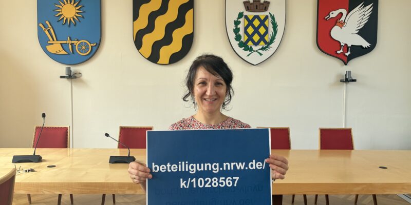 Eine Frau steht in einem Rathaussaal und hält ein Schild in der Hand. Darauf zu lesen: beteiligung.nrw.de