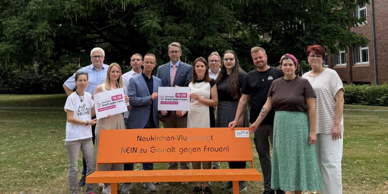Menschen stehen hinter einer Bank. Die Bank trägt die Aufschrift: Neukirchen-Vluyn sagt NEIN zu Gewalt gegen Frauen
