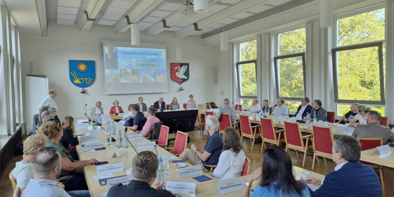 Viele Menschen sitzen in einem Saal an langen Tischen und schauen nach vorn auf eine Leinwand