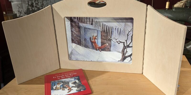 Ein Bilderbuch liegt auf einem Holztisch. Dahinter eine Illustration in einem Bildaufsteller aus Holz.