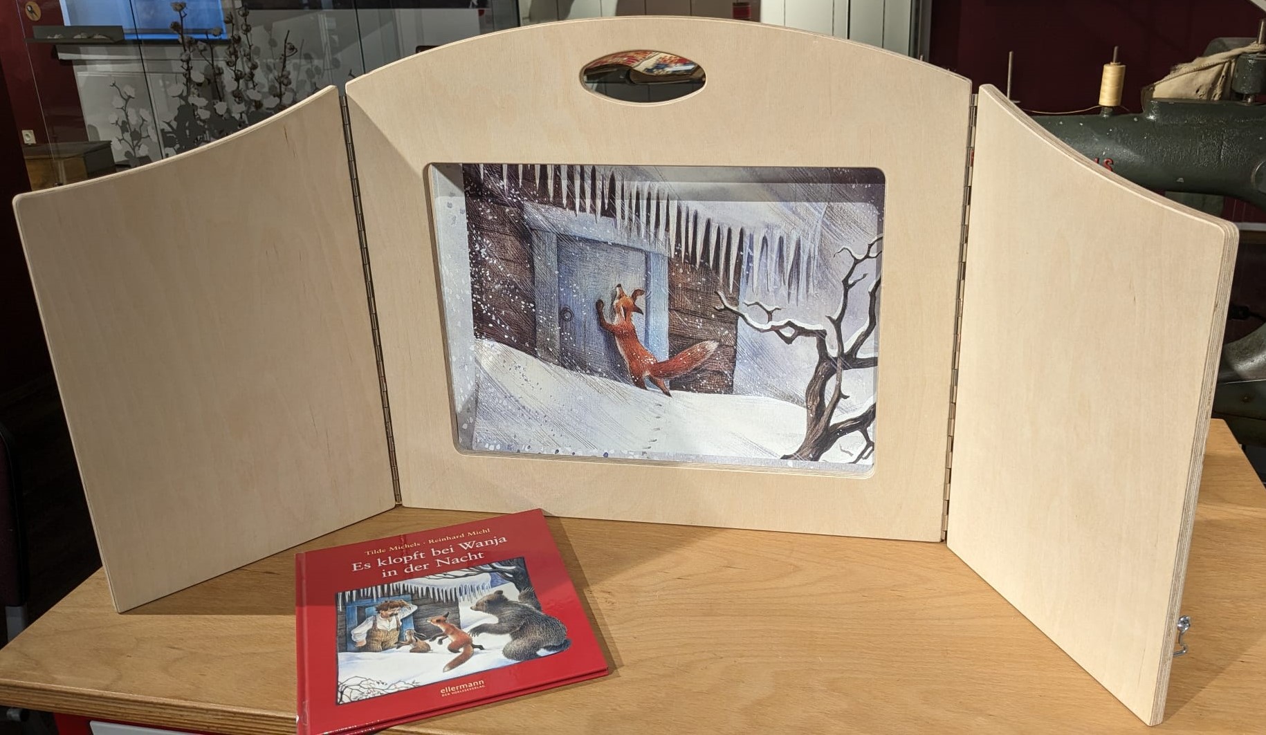 Ein Bilderbuch liegt auf einem Holztisch. Dahinter eine Illustration in einem Bildaufsteller aus Holz.