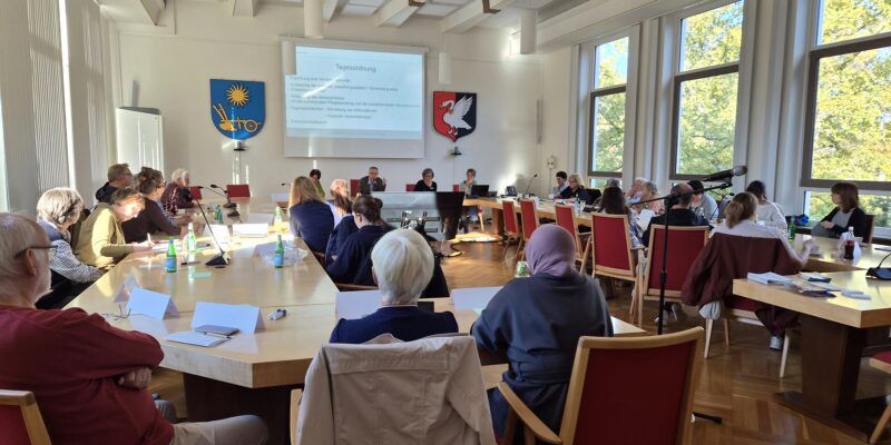 Menschen sitzen in einem Saal an Tischen zusammen.